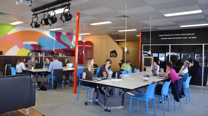 Open-plan coworking space with colourful mural walls, stage lighting rig, and people working at shared tables