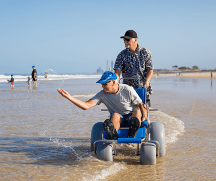 Man being pushed through shallow waves in a blue beach wheelchair at Leighton Beach, bookable on SpacetoCo