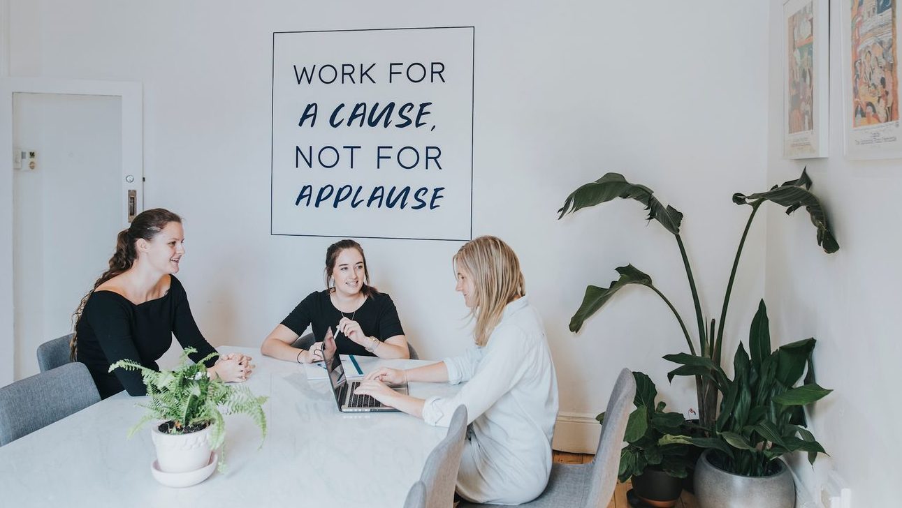 Three people sitting around a table with a laptop and potted plants, with a "work for a cause, not for applause" sign on the wall behind them