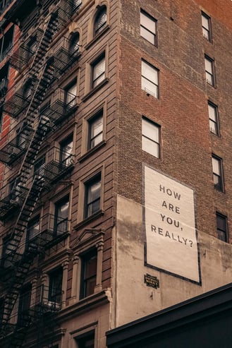 Brick apartment building with a fire escape and a painted sign reading "how are you, really?"